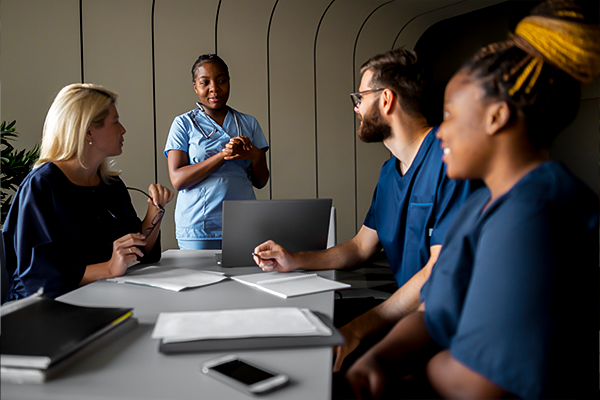 A diverse group of healthcare professionals in scrubs is engaged in a team meeting around a table with laptops and papers, indicative of a discussion about workplace safety or workers' compensation in healthcare.