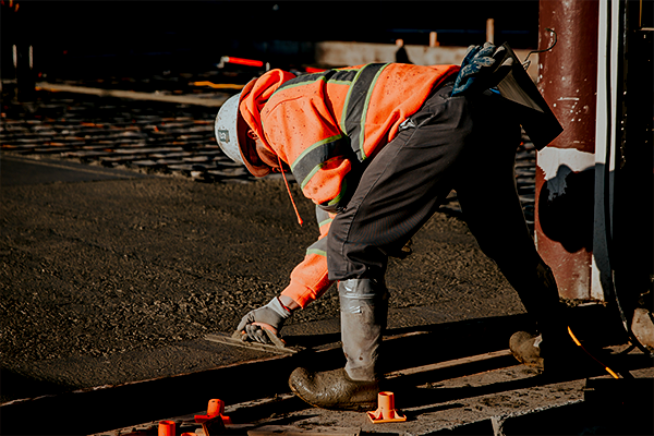 A construction worker in a reflective orange vest and safety helmet is bent over working on asphalt, highlighting the physical nature of the job relevant to workers' compensation
