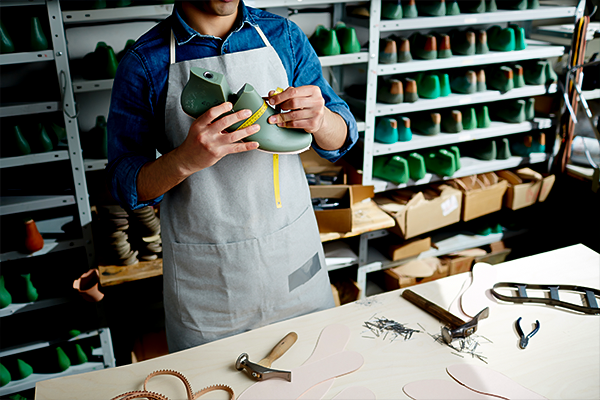 A craftsman in an apron measures a piece of green material for shoemaking in a workshop, surrounded by tools and shelves of shoes, representing small business labor relevant to workers' compensation insurance.