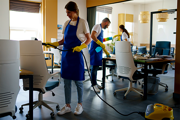 Alt text: "A team of cleaners in a bright office setting, with a woman in the foreground wiping a desk while wearing a blue apron and yellow gloves, indicative of the types of employees that might be covered by workers' compensation insurance for cleaning services.