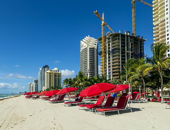 Red beach umbrellas and lounge chairs on the sand with towering skyscrapers and construction cranes in the background, possibly in a Florida coastal city, evoking thoughts of work and leisure balance and worker protections in such environments.