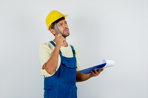 A construction worker in a yellow hard hat and blue overalls holds blueprints and appears thoughtful, possibly considering project details or workers' compensation matters.