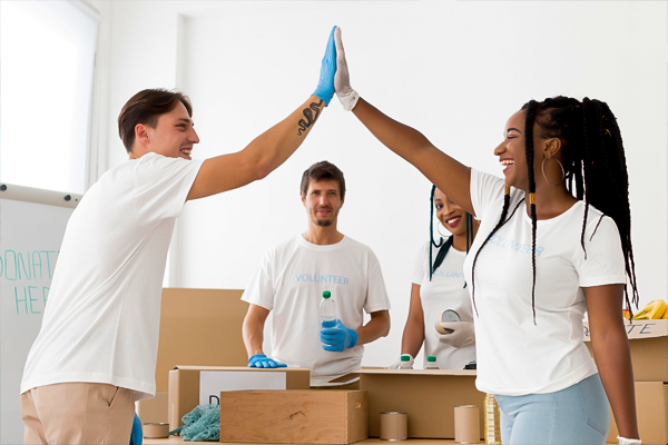 Four cheerful volunteers wearing 'VOLUNTEER' t-shirts are giving a high-five in a bright room, with cardboard boxes and cleaning supplies around, possibly reflecting team spirit and the importance of workers' compensation in non-profit settings.