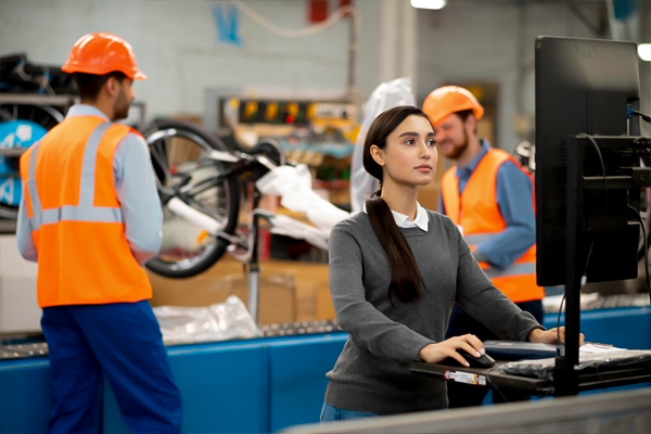 Alt text: "A focused female worker in a warehouse, standing in the foreground at a computer station, with male colleagues in safety vests and helmets in the background, illustrating a workplace environment relevant to workers' compensation insurance topics.